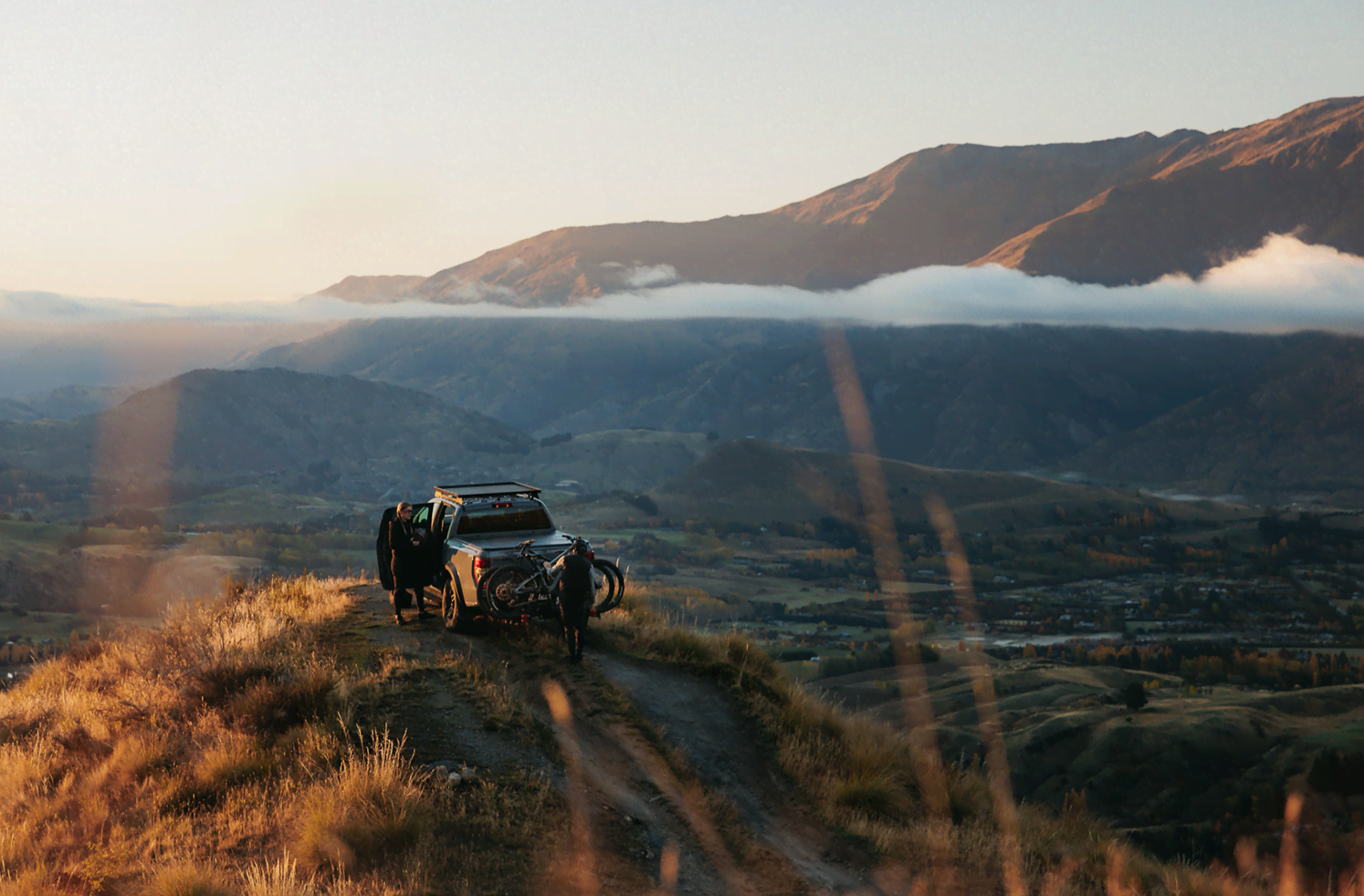 A ute with a bike rack on the back sits atop a mountain as a mountain biker unloads his rack