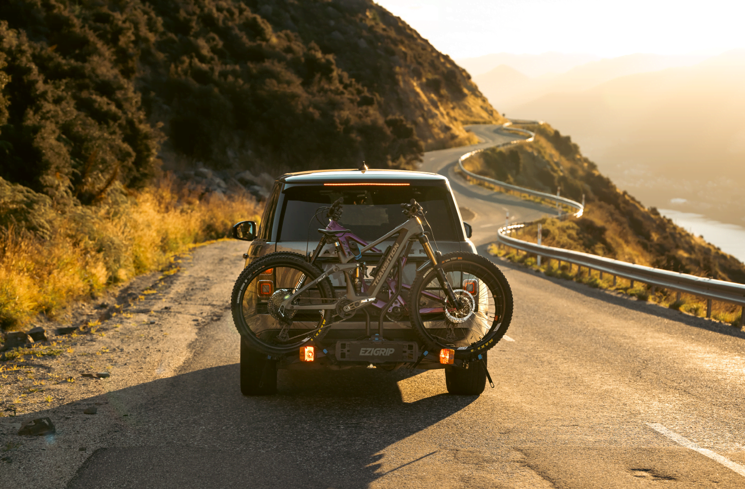 A bike rack is mounted to the back of a vehicle and driving into the sunset
