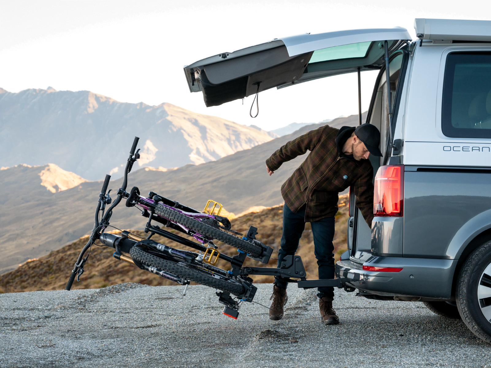 A man is reaching into the back of a van with the tailgate open. The Ezigrip E-Rack 2 Pro bike rack is attached to the back of the van and tilted away from the van while holding two ebikes.