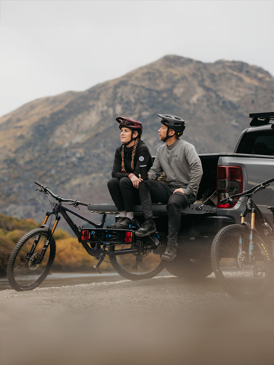 Two mtb riders are sitting on the back of a tailgate of a ute. They rest their feet on an Enduro 2 bike rack.