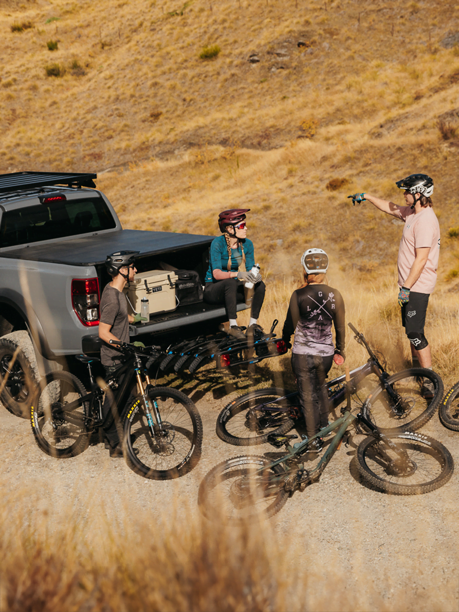 A group of mtb riders sit around a ute with an Enduro 4 bike rack mounted to the back. The rack is folded down and the tailgate of the ute is open.