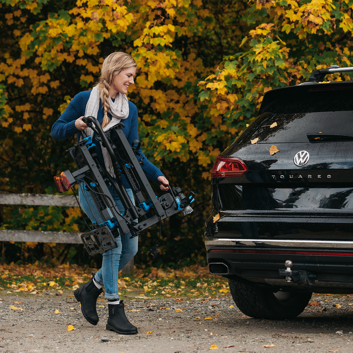 A woman is carrying an E-Rack 2 Bike Rack to a towball on the back of an SUV. The bike rack is folded and close to her chest as she carries it using the handle.