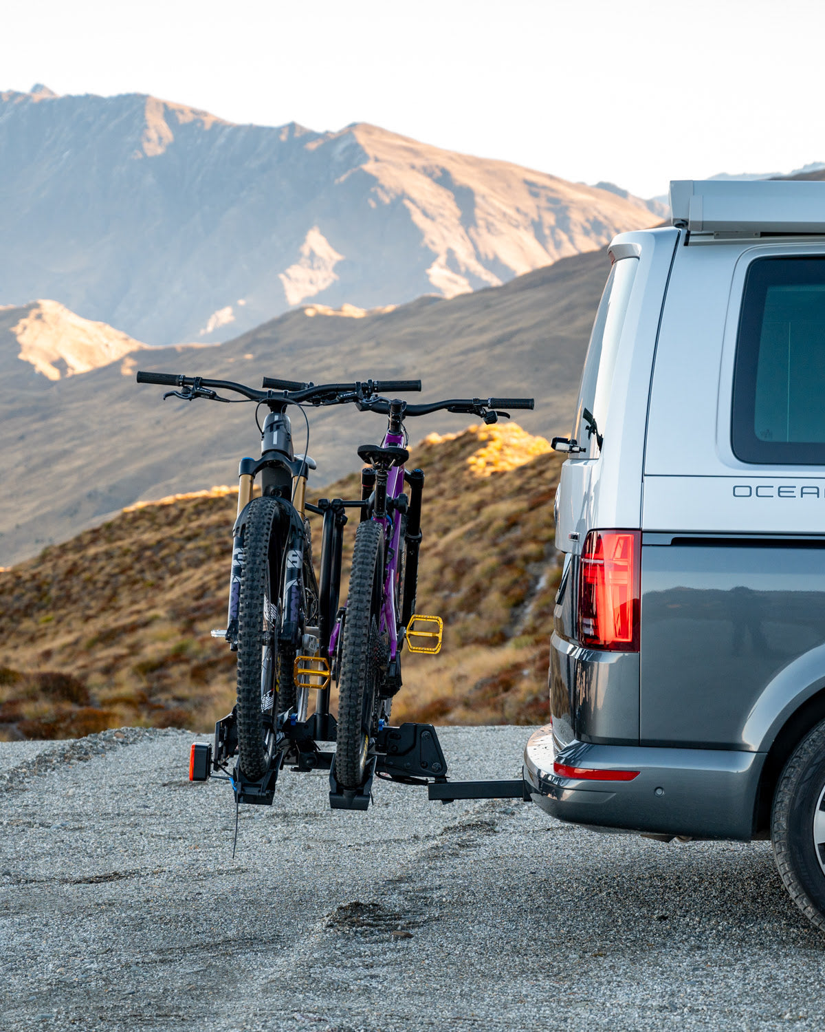 Two electric bikes are sitting snugly on the Ezigrip E-Rack 2 Pro hitch mounted ebike rack. The rack is mounted to the hitch of a van and in the background you can see the mountains of south New Zealand.