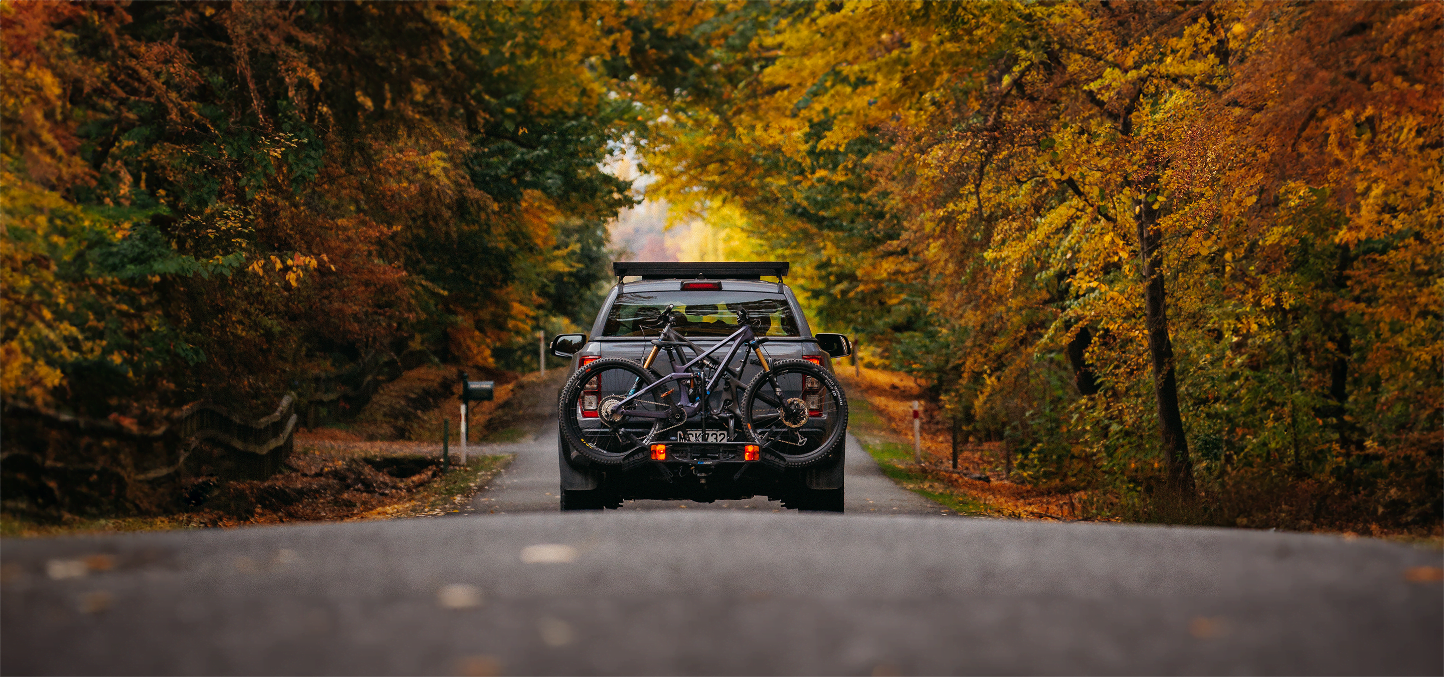 A truck drives down a country road with the forest either side. On the back of the truck is an Ezigrip Enduro 2 bike rack hauling two mountain bikes.