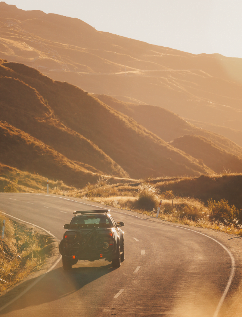 A vehicle with a bike rack installed on the back is driving up a winding mountain road with the Coronet Peak in the background