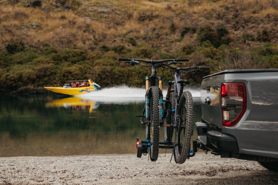 A bike rack with two mtbs on the rack attached to a ute is visible in front of a lake as a jetboat cruises by on the river behind it