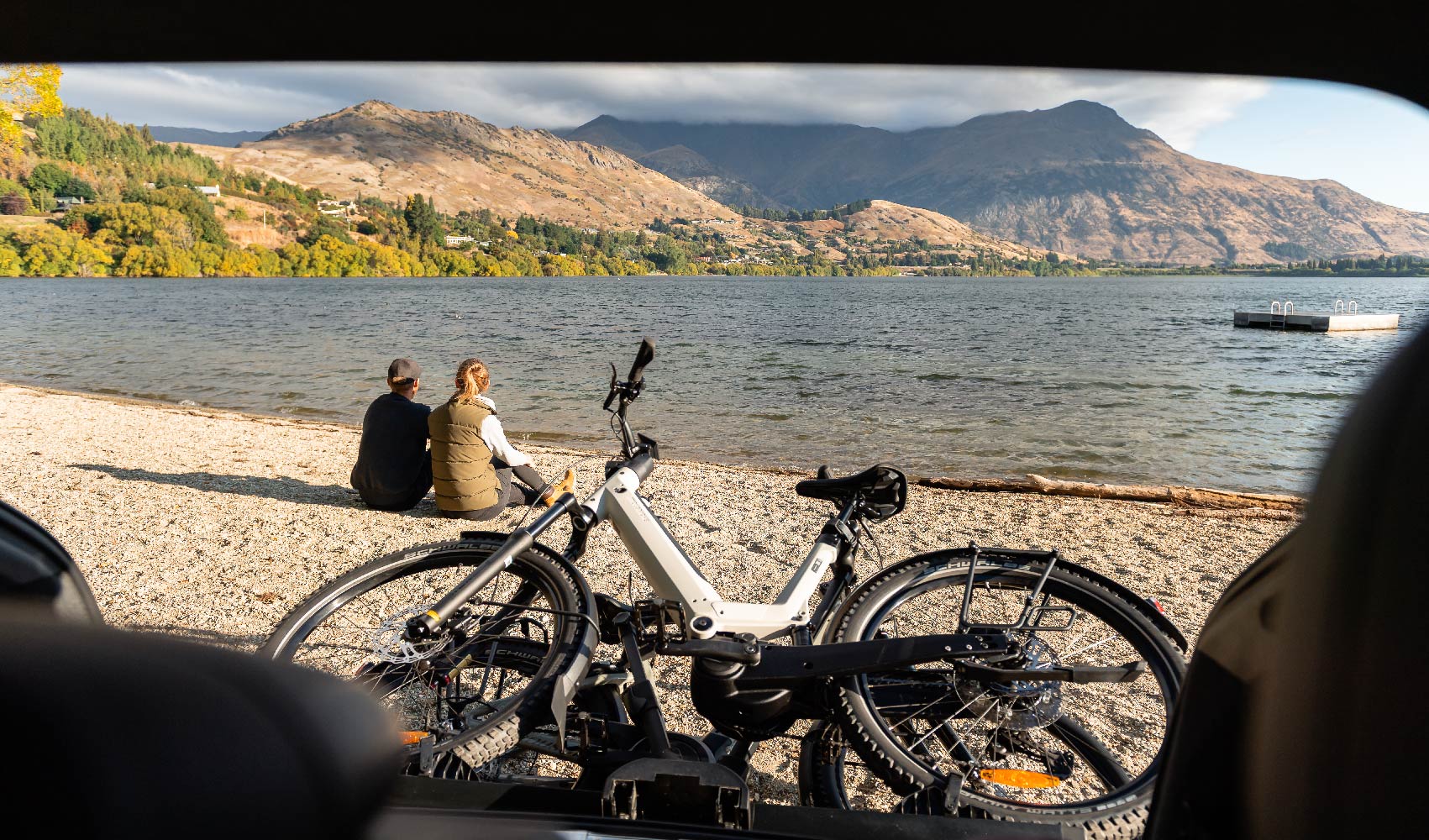 A man and a woman sit looking at a lake while their mountain bikes are tilted on a bike rack on the back of a vehicle