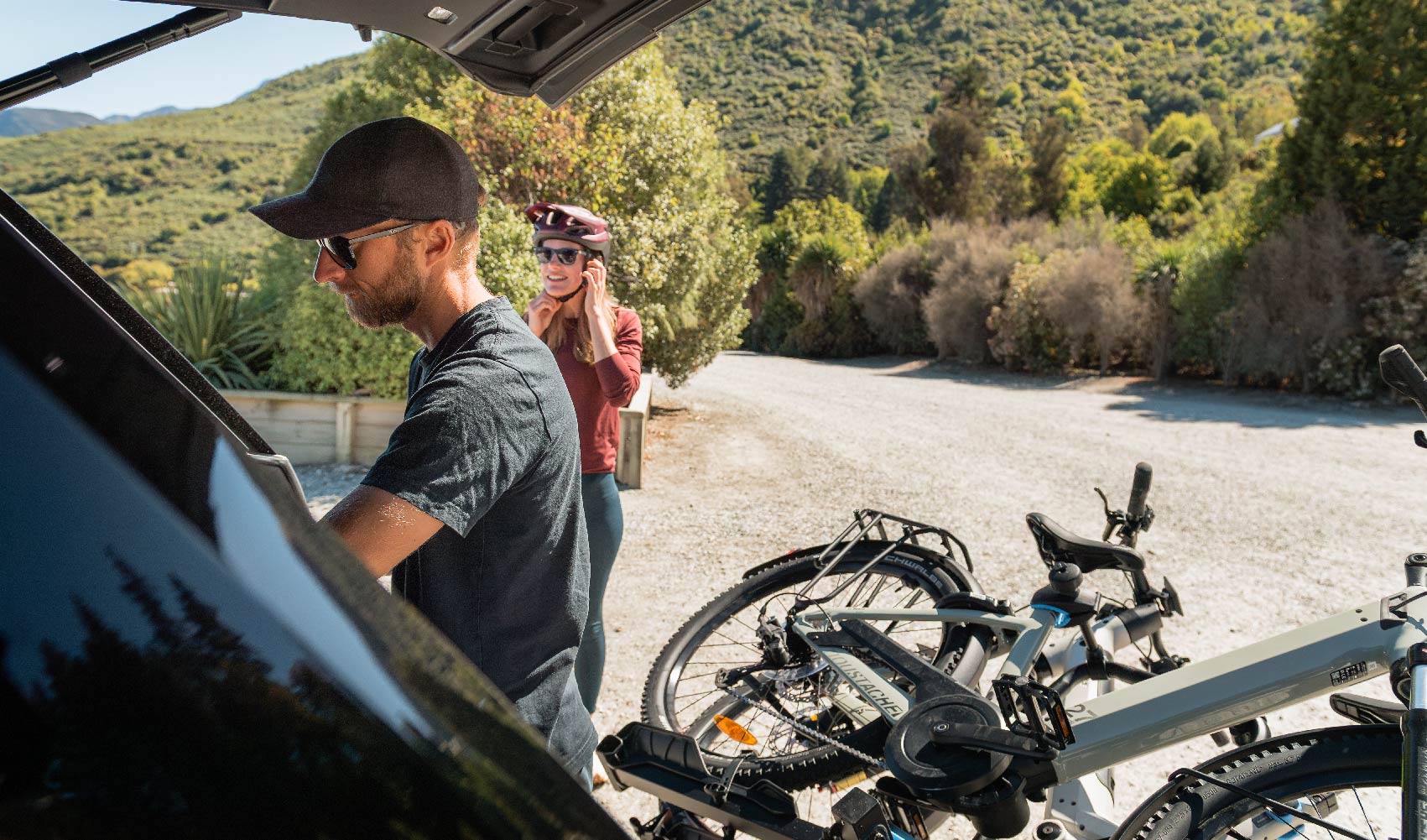 A man and a woman prepare for a bike ride. Two ebikes are on a bike rack, tilted away from the tailgate