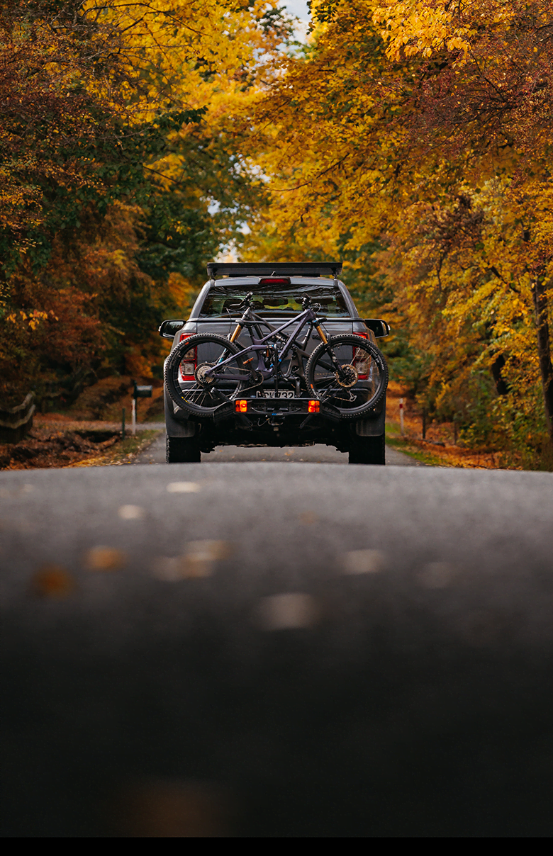 A truck drives down a country road with the forest either side. On the back of the truck is an Ezigrip Enduro 2 bike rack hauling two mountain bikes.