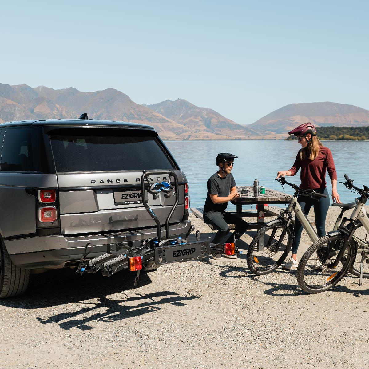 A man and a woman prepare for a bike ride by a lake. a bike rack is clearly visible, mounted to a range rover