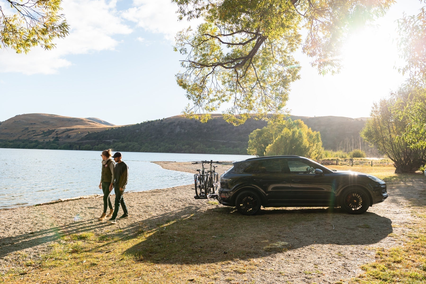 A man and a woman walk along a lake. A vehicle is nearby with an Ezigrip E-Volve 2 bike rack on the back and two electric bikes strapped to the rack