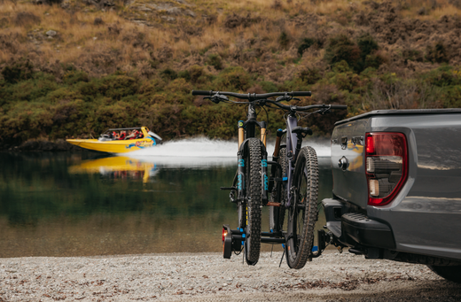 An Ezigrip Enduro 2 bike rack is mounted to the towball of a ute with two mountain bikes strapped into the bike rack. A jet boat speeds by in the background.