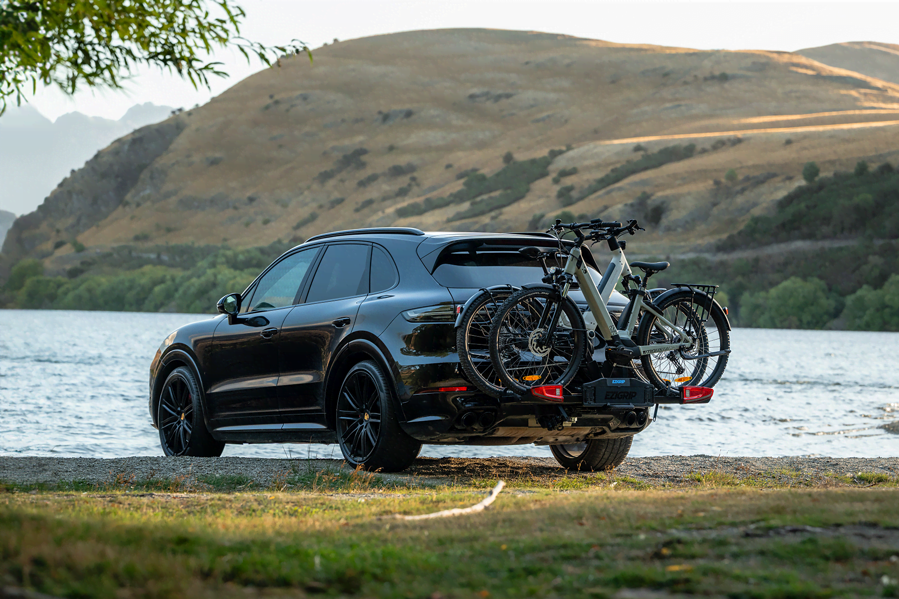 A Porsche SUV sits by a lake with two step-through mountain bikes attached to the Ezigrip E-Volve 2 bike rack