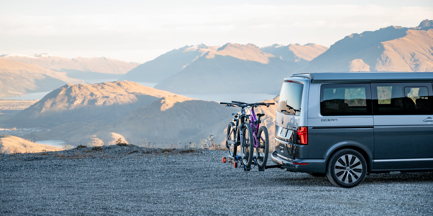 A van with a bike rack attached to the back and loaded with two electric bikes sits in the foreground with the sweeping New Zealand mountains in the background