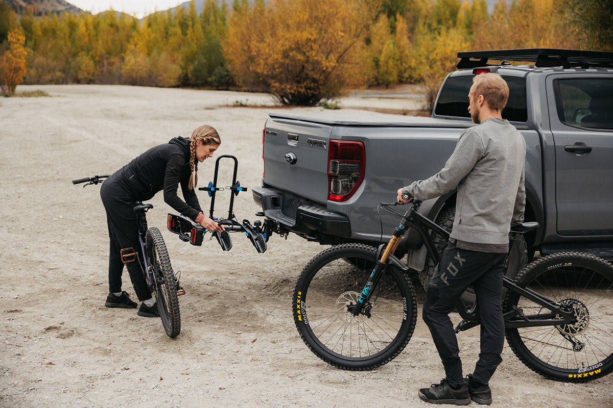 A woman is preparing an Ezigrip Enduro 2 bike rack for loading a bike. The bike rack is mounted to a tow ball of a vehicle.