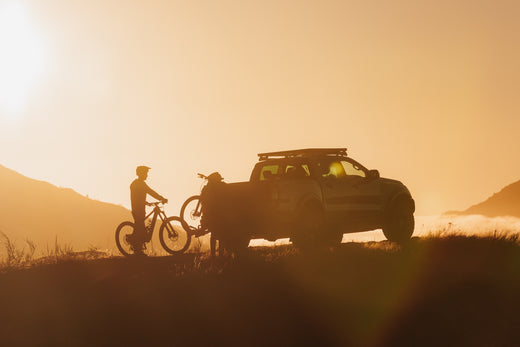 The silhouette of a truck with a bike rack mounted on the back and two riders wheeling their bikes into position.