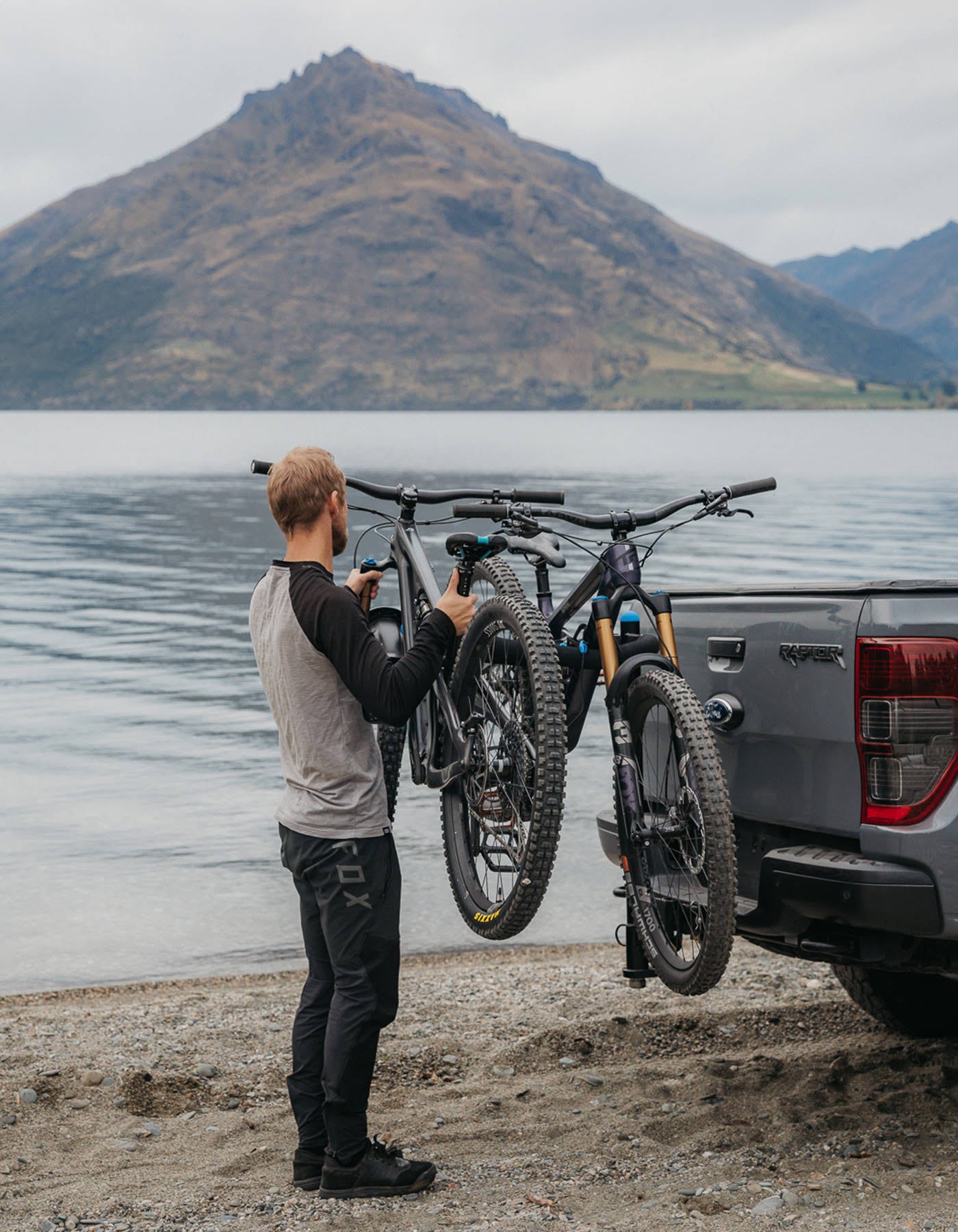 A man is loading a mountain bike onto an Ezigrip Advantage 2 bike rack that is attached to a Ford Ranger. There is mountains and a lake behind him.