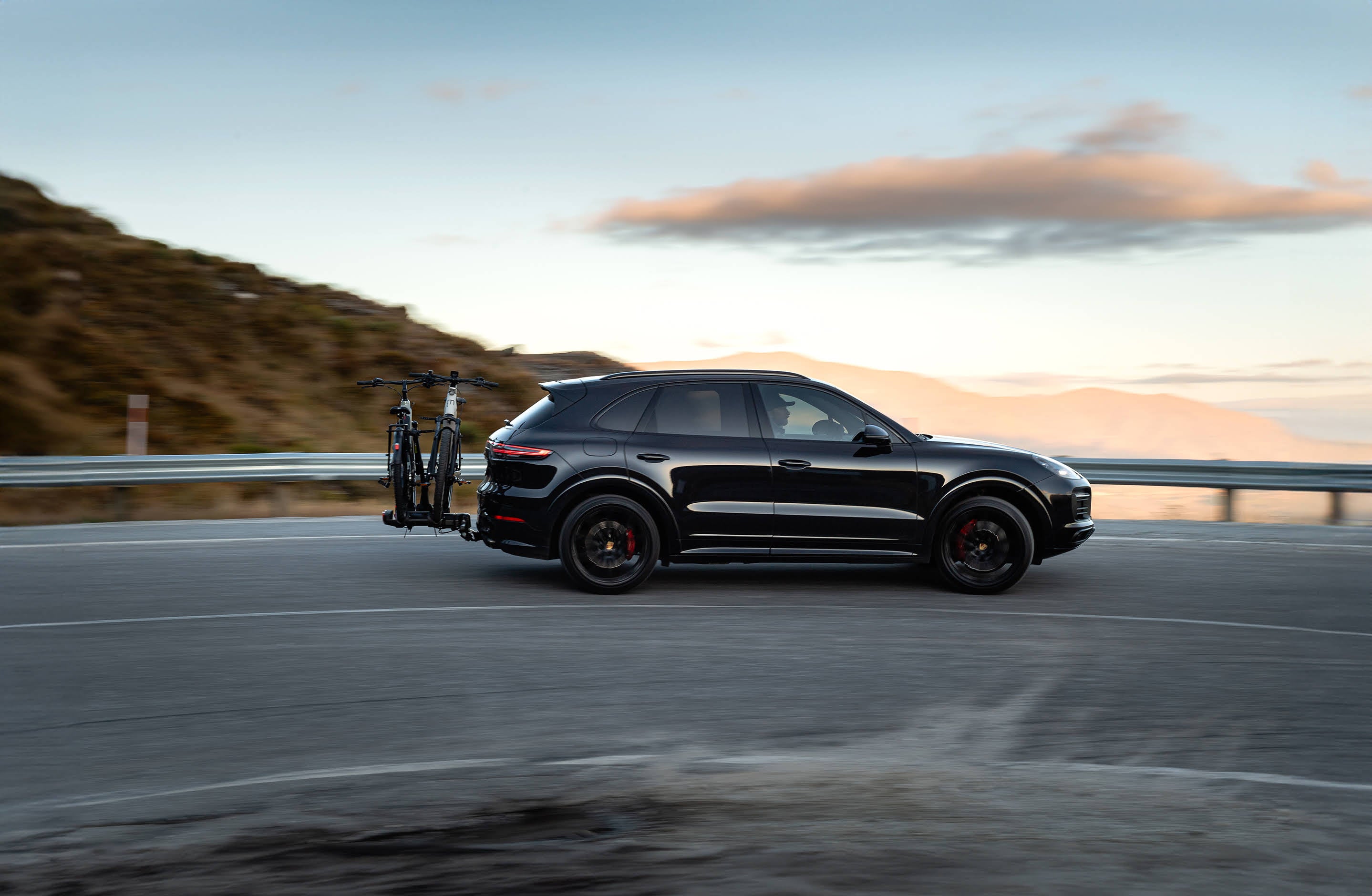 A Porsche SUV drives around a mountain road with an Ezigrip E-Volve 2 bike rack on the back carrying two electric bikes
