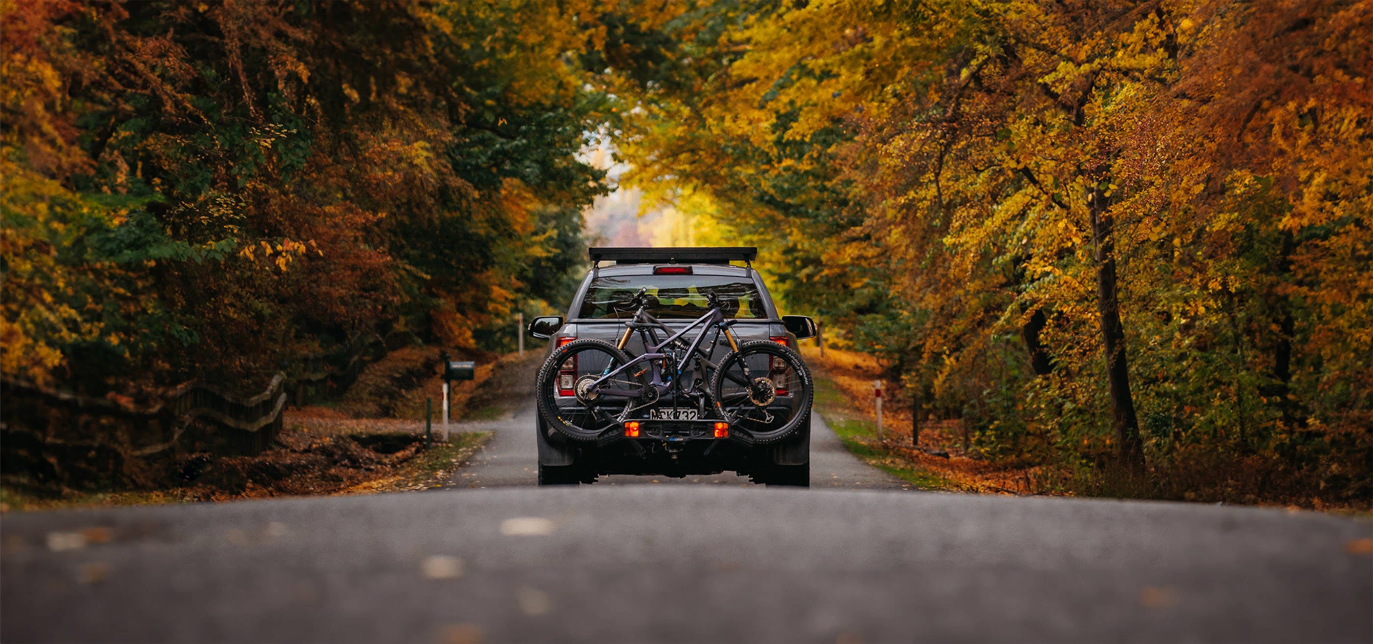 A truck drives down a country road with the forest either side. On the back of the truck is an Ezigrip Enduro 2 bike rack hauling two mountain bikes.