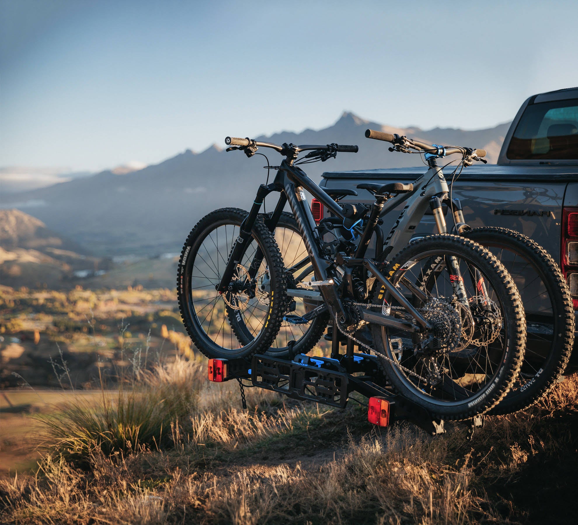 An E-Rack 2 bike rack with two electric bikes mounted to the rack is visible on the back of a ute with a sweeping mountain in the background