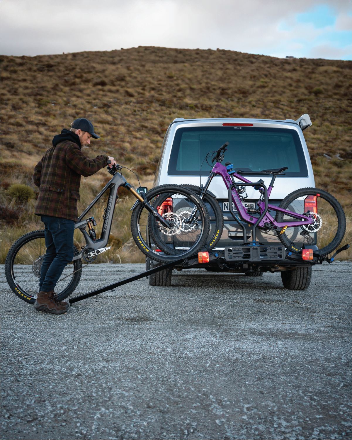 A man is rolling an electric bike up a ramp onto the Ezigrip E-Rack 2 Pro ebike rack which is mounted to the back of a van. A second ebike is already loaded on the bike rack.