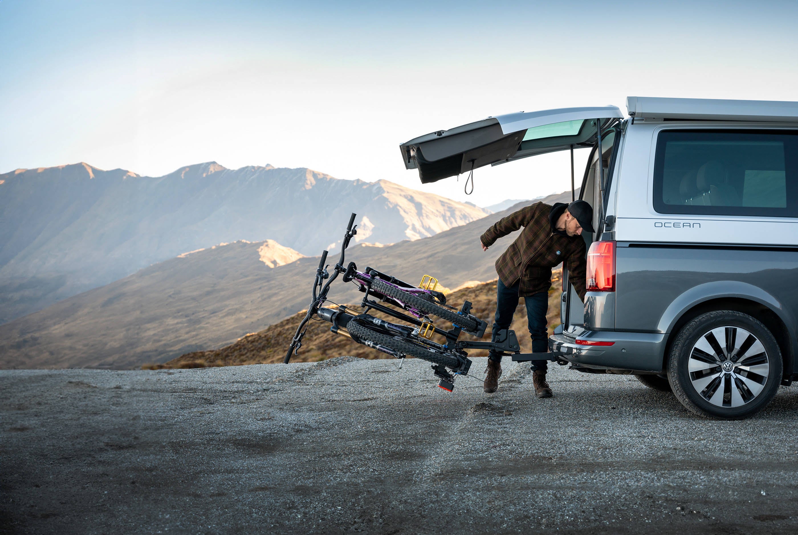 A man reaches into the tailgate of the van with a bike rack on the back. The bike rack is tilted away from the van with the bikes on the rack.