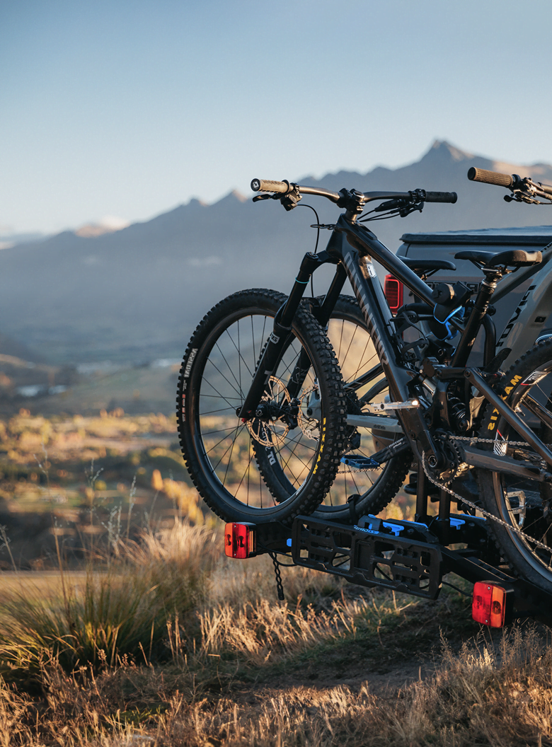 An E-Rack 2 bike rack with two electric bikes mounted to the rack is visible on the back of a ute with a sweeping mountain in the background