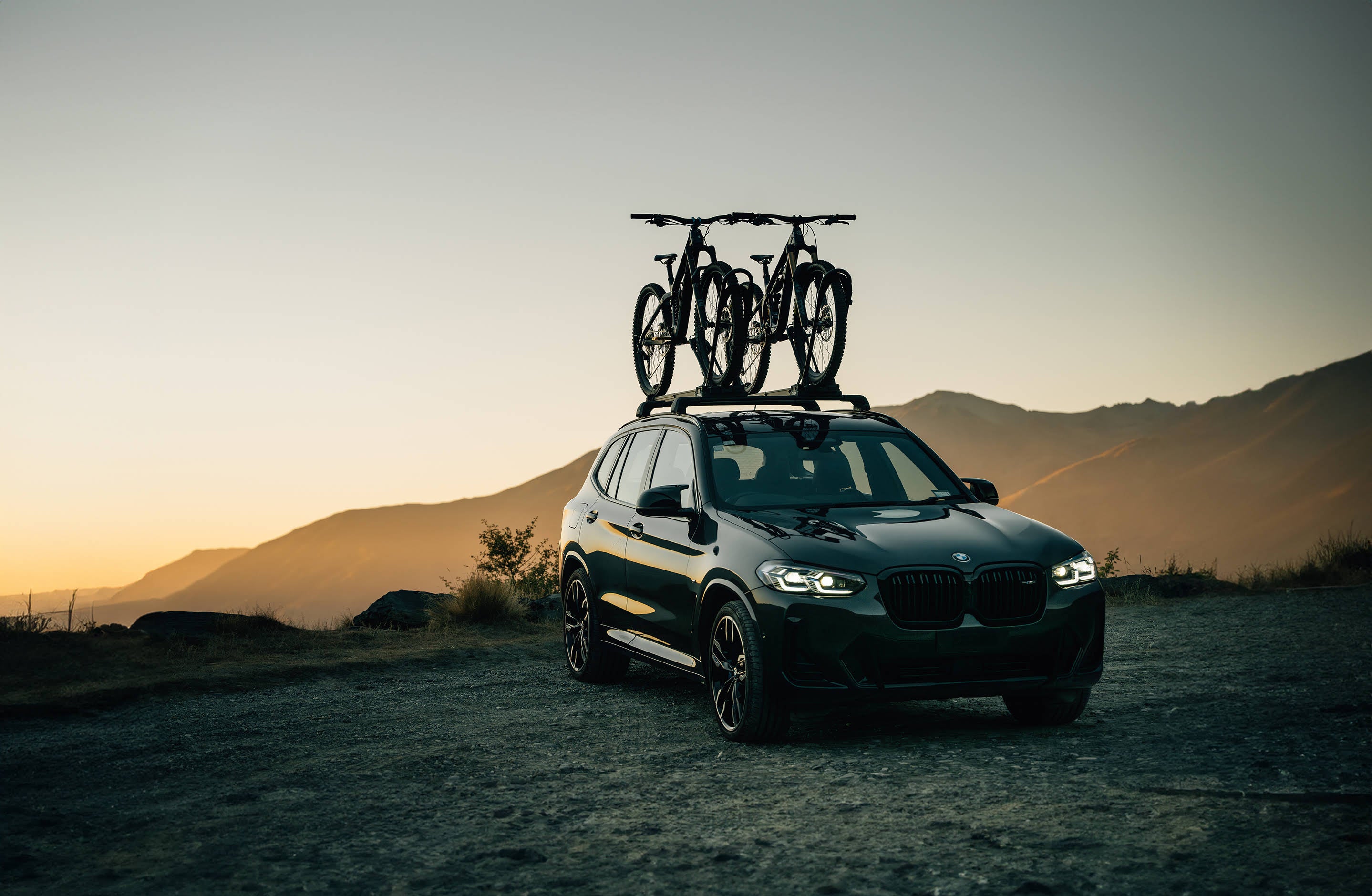 A sleek BMW SUV sits atop a mountain as the sun rises with two mountain bikes on the roof, secured in place by the Ezigrip Uplift 1 bike rack